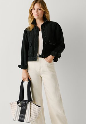 Nuori nainen, jolla on musta lyhyt takki ja vaaleat housut, pitää raidallista ostoskassia mustilla kahvoilla yksivärisen taustan edessä.