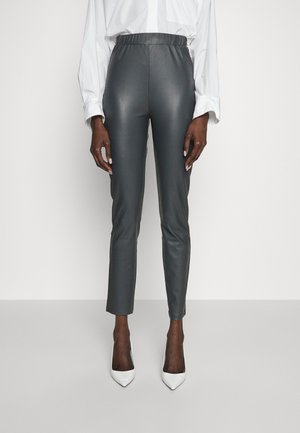 Kobieta w ciemnoszarych, skórzanych spodniach z wysokim stanem i białych, z pointed-toe szpilkach, ubrana w białą bluzkę z długim rękawem na jednolitym tle.