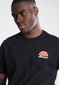 Black cotton t-shirt featuring a small red, orange, and white logo on the left chest, crew neck design, and short sleeves. Simple texture.