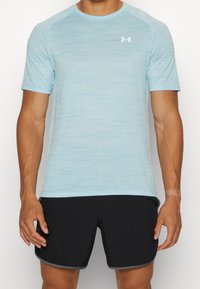 Camiseta deportiva de color azul claro, que absorbe la humedad, con mangas cortas, tejido texturizado y un logo blanco en el lado izquierdo del pecho, combinada con pantalones cortos negros.