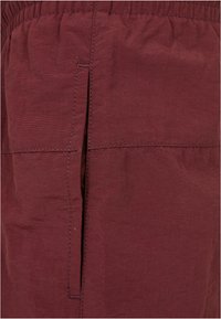 Shorts color bordeaux realizzati in tessuto leggero, dotati di una cintura elastica, tasca laterale e dettagli di cucitura a vista. Texture liscia.