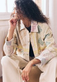 Gesteppte Jacke mit mehrfarbigen Blumen- und geometrischen Mustern. Baumwollmischgewebe, Knopfverschluss und lockere Passform. Cremefarbener Hintergrund.