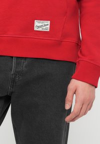 Κόκκινο φούτερ με λευκή ραμμένη ετικέτα κοντά στο τελείωμα και στους καρπούς, φορεμένο με μαύρο τζιν, κοντινή λήψη κάτω μέρους κορμού και δεξιού χεριού.