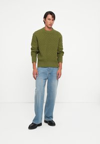 Pulover verde tricotat cu un model texturat în formă de romb, asociat cu blugi largi de culoare albastru deschis și bocanci negri masivi. Design simplu și contemporan.