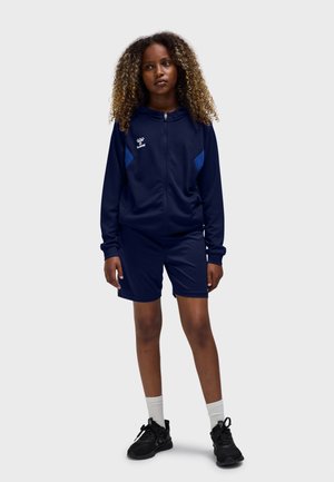 Marineblåt atletisk træningssæt bestående af en lynlåsjakke med blå accenter og matchende shorts, kombineret med sorte sneakers og hvide sokker.