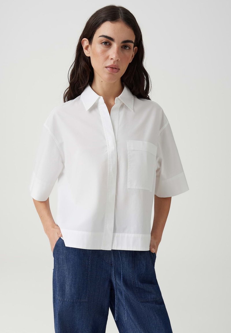 Woman with long dark hair wearing a short-sleeve white collared shirt and dark blue wide-leg pants, standing with hands in pockets.