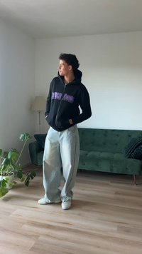 Felpa nera con zip e testo grafico viola, jeans larghi di un azzurro chiaro e sneakers bianche. In piedi in una stanza con divano verde e pianta.