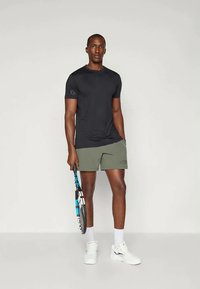 Svart kortermet treningsskjorte kombinert med olivengrønne shorts, holder en tenniskølle. Joggeskoene er hvite; synlige logoer på klærne.