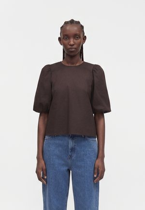 Femme aux cheveux tressés portant un haut sombre à manches bouffantes et un jean bleu taille haute, debout devant un fond blanc uni.
