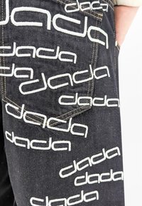 Pantalones de mezclilla negra con impresiones repetidas del logotipo blanco "dada", con costuras visibles y un detalle de bolsillo trasero.
