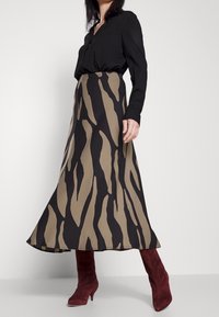 Maxiröcke in Beige mit schwarzen abstrakten Streifen, aus glattem Stoff gefertigt. Kombiniert mit einer schwarzen Bluse und burgunderfarbenen Wildlederschnürstiefeln.
