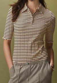 Beżowa koszulka polo z krótkim rękawem w cienkie czarne paski. Posiada kołnierzyk i falbaniaste krawędzie rękawów, w połączeniu z szarymi spodniami.