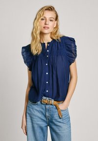 Blusa blu navy con maniche corte a sbuffo, chiusura con bottoni e dettagli arricciati. Indossata con jeans in denim blu chiaro e una cintura marrone.