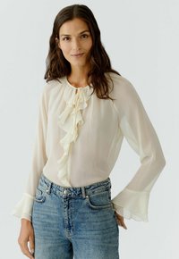 Cremefarbene Bluse mit rüschengeschmückter Vorderseite, langärmelige Ärmel mit voluminösen Rüschenbündchen, kombiniert mit hochgeschnittenen blauen Jeans. Leichter, strukturierten Stoff.