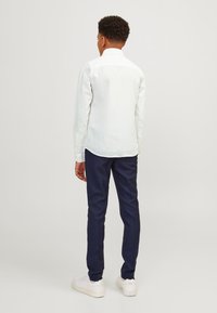 Camicia bianca a maniche lunghe con colletto a punta, abbinata a pantaloni blu navy e sneakers bianche. La camicia ha una texture liscia e un orlo curvo.