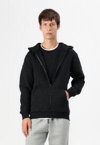 Sudadera con cremallera negra, con capucha, hecha de tela suave. Cuenta con bolsillos frontales y puños acanalados, llevada sobre una camiseta negra y pantalones de chándal grises.