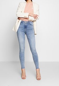 Jeans ajustados de color azul claro con una cintura alta, combinados con una blusa rosa pálido y un blazer crema. Sandalias de tacón transparentes completan el look.