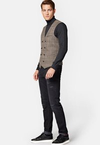 Brązowa kamizelka w kratę na czarnym golfie, zestawiona z czarnymi zniszczonymi dżinsami i czarnymi sneakersami. Teksturowany materiał i dopasowany krój.