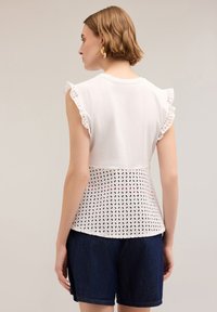 Top blanco con mangas acampanadas, que presenta un patrón de encaje texturizado en la cintura. Combinado con pantalones cortos de mezclilla oscura.