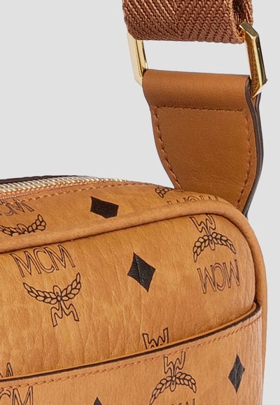 Sangle en cuir marron avec boucle en or attachée à un sac beige arborant le logo MCM noir et un motif en diamant sur du cuir texturé.