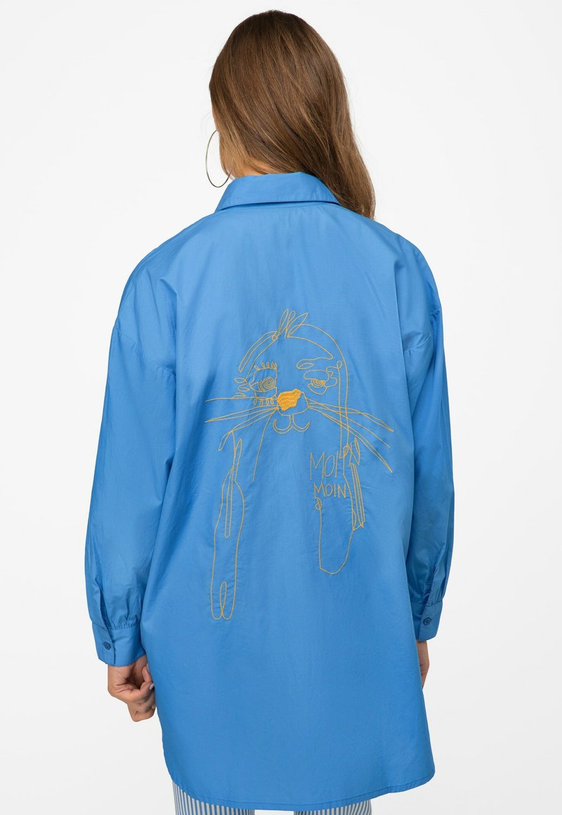 Light blue oversized shirt with long sleeves. Features gold embroidery of a cartoon character on the back. Smooth texture with buttoned cuffs.