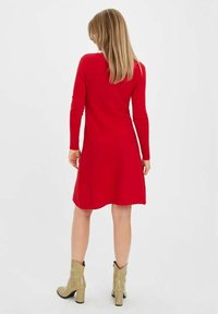 Robe tricotée rouge à manches longues et à ourlet évasé, présentant une texture lisse. Portée avec des bottines beiges à texture.