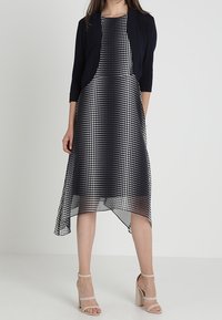 Frau trägt ein schwarz-weißes Polkadot-Midikleid mit asymmetrischem Saum und einer schwarzen, kurzen Strickjacke, kombiniert mit beigen High-Heels.