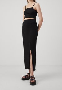 Femme portant un débardeur court noir, une jupe midi noire taille haute avec une fente latérale, et des sandales noires à boucle, debout devant un fond uni.