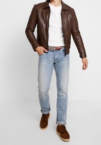 Brązowa skórzana kurtka z kołnierzem, biała teksturowana koszula, jasnoniebieskie dżinsy oraz brązowe zamszowe buty z jasną podeszwą.