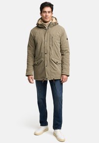 Olivově zelená manšestrová bunda s podšitou kapucí, předními kapsami a zipovou kapsou na hrudi. Stylizováno s modrými džínami a světlými botami.