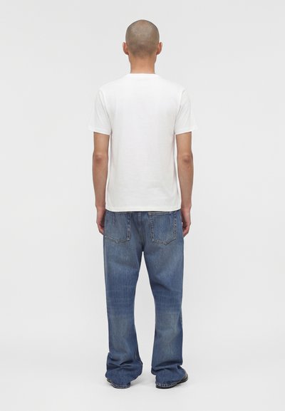 Άνδρας με ξυρισμένο κεφάλι στέκεται πλάτη, φοράει ένα απλό λευκό κοντομάνικο μπλουζάκι και φαρδιά μπλε τζιν μπροστά σε ουδέτερο φόντο.