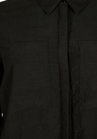 Camicia nera con bottoni realizzata in un tessuto testurizzato, dotata di colletto e due tasche frontali. Il design è minimalista e sartoriale.