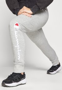 Grå joggebukser med en hvit brodert "Champion" logo langs siden og en rød detalj. Kombinert med svarte treningssko.