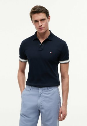 Uomo che indossa una polo blu navy con maniche a righe bianche e pantaloni azzurri, in piedi con le mani in tasca davanti a uno sfondo uniforme.