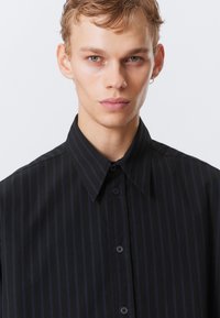 Musta kauluspaita, jossa on pystysuuntaisia tummansinisia raitoja. Paidassa on napitus edessä ja sileä kangas. Yksinkertainen muotoilu, ilman koristeita.