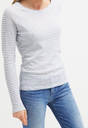 Femme portant une chemise blanche cintrée à manches longues avec des rayures horizontales grises et un jean bleu, debout devant un fond blanc uni.