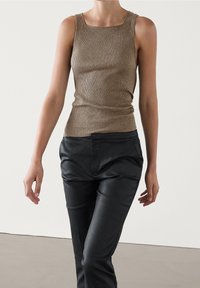 Personne portant un débardeur marron côtelé sans manches et un pantalon noir taille haute, debout les bras détendus contre un fond uni.