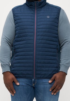 Homme portant une veste matelassée bleu marine avec fermeture éclair sur une chemise bleu clair à manches longues et un jean bleu, les mains reposant sur les côtés.