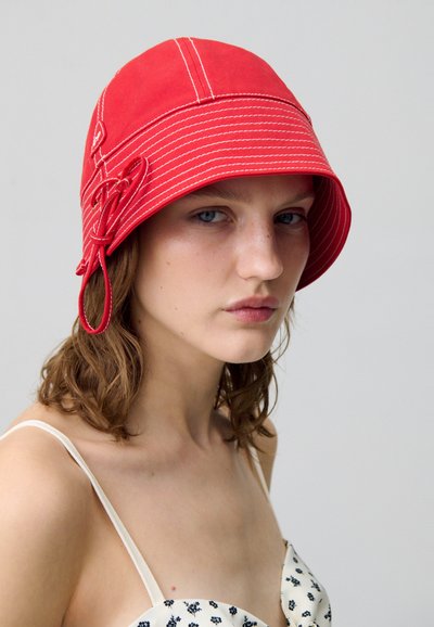 Cappello a secchiello rosso in tessuto con cuciture bianche, dalla forma rotonda e con un legaccio decorativo sul lato.