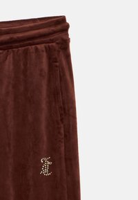 Smeđe velur hlače s mekim teksturom, s bočnim džepovima i detaljem s cirkonima u obliku loga. Elastična traka je glatka i ravna.