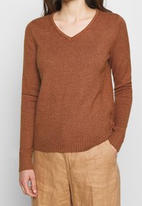 Brun, långärmad tröja i ett strukturerat material. Har en V-ringning och ribbad nederkant, i kombination med ljusa, löst sittande byxor.