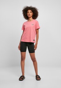 Tricou din bumbac roz, cu guler rotund și mâneci scurte, având un mic patch cu logo și asortat cu pantaloni scurți de ciclism negri și sandale.