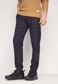 Marinblå rutiga byxor med blå och bruna linjer, som har en slim fit. Kombinerat med svarta sportskor; en strukturerad brun stickad tröja ovanför.