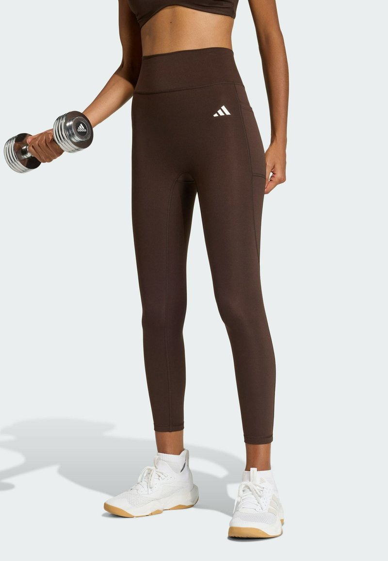 Leggings marron taille haute avec une texture lisse, arborant un logo Adidas blanc et des coutures détaillées, assortis à des chaussures de sport blanches.