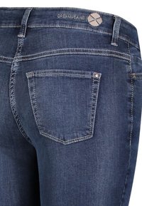 Dunkelblaue Jeans aus Denim mit glatter Textur, ausgestattet mit einer Gesäßtasche und einem gestickten Logo am Bund. Klassisches Fünf-Taschen-Design.