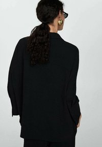Chemise noire surdimensionnée à manches longues, en tissu texturé, avec des accents de boutons. Présente une coupe droite et un détail de yoke au dos.