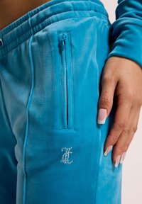 Blaue Velours-Sweatpants mit einem Reißverschlussfach, das ein kleines Kristall-Logo am Oberschenkel trägt. Glatte Textur und elastischer Bund sind sichtbar.