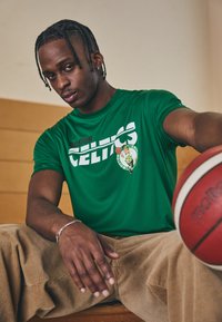Zöld Boston Celtics póló fehér szöveggel és logóval, bézs kordbársony nadrággal párosítva. A sárga részletekkel rendelkező kosárlabda a foregroundban pihen.
