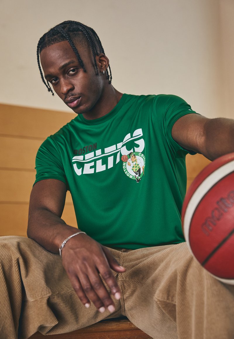 Zöld Boston Celtics póló fehér szöveggel és logóval, bézs kordbársony nadrággal párosítva. A sárga részletekkel rendelkező kosárlabda a foregroundban pihen.