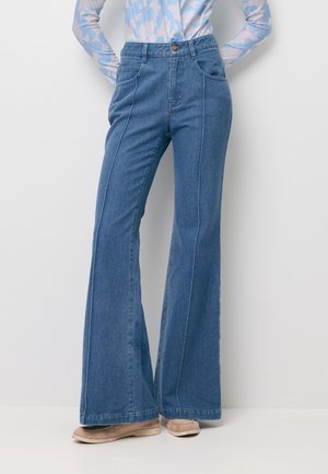 Vrouw die een blauwe jeans met hoge taille en brede pijpen draagt, beige schoenen en een lichtblauw patroon lang shirt, staat met handen in de zij.